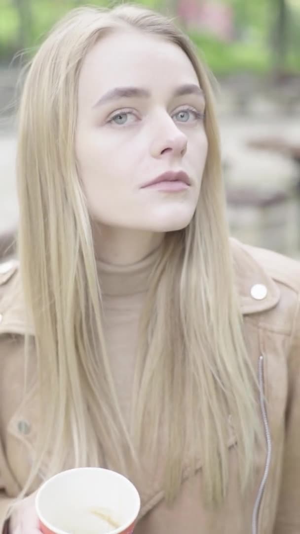 Vertical video of a young skinny girl in the park. The girl is drinking coffee. — Stock Video