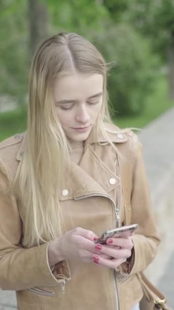 Вертикальное видео молодой худой девушки в парке. Девушка с мобильным телефоном. — стоковое видео