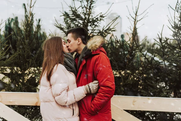 A young couple kissing in the winter against the background of the Christmas tree market in the city. love, winter holidays and people concept — Stock Photo, Image