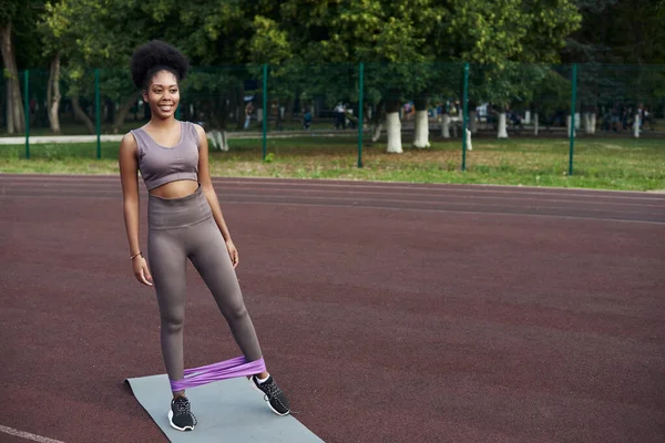 Sötét bőrű lány ruganyos fitneszgyűrűvel rázza az alsó lábait. Szabadtéri sportok — Stock Fotó