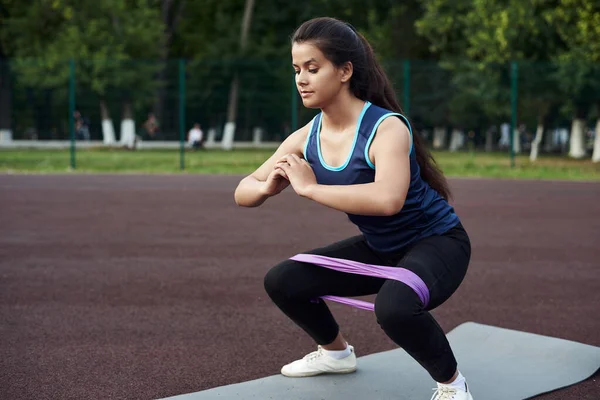 Ung kvinna går in för sport med hjälp av ett elastiskt band för fitness. En vacker sportig flicka skakar på benen med en expander — Stockfoto