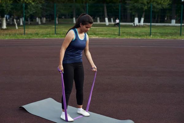 Een mooie vrouw schudt de spieren en spieren in haar armen met behulp van een expander en fitness elastiekjes. Schattig meisje gaat in voor sport op straat — Stockfoto