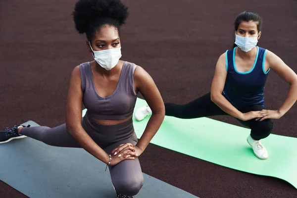 Twee verschillende etnische meisjes strekken zich uit, strekken hun spieren uit alvorens buiten te trainen met een beschermend masker. Preventie van coronavirus — Stockfoto