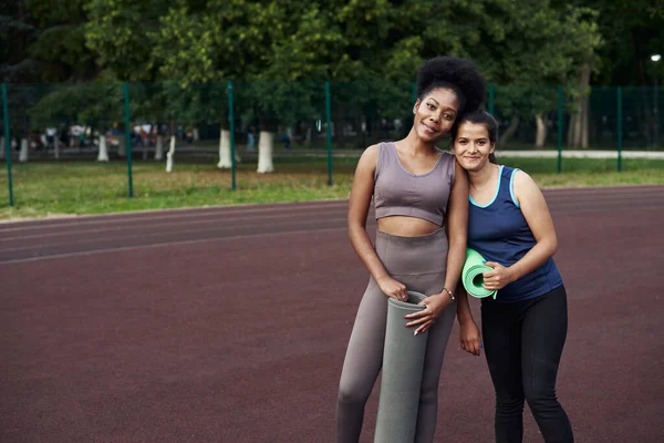 Due donne sportive tengono in mano un tappetino da yoga e vanno ad allenarsi allo stadio per strada. Bellissimi modelli durante l'allenamento all'aperto — Foto Stock