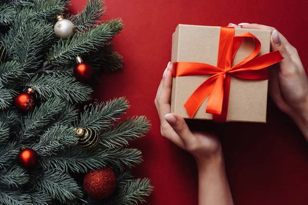 Weibliche Hände halten Weihnachtsgeschenk auf rotem Hintergrund mit Tannenzweigen und Kugeln. Xmas und Happy New Year Karte. Flache Lage, Draufsicht — Stockfoto