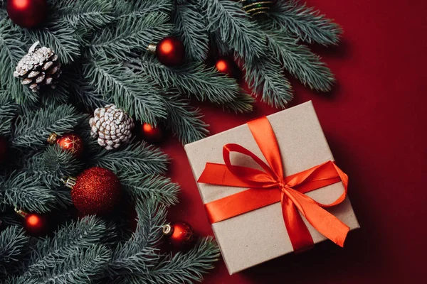 Regalo artesanal con una cinta de satén rojo junto a las ramas y bolas del árbol de Navidad, juguetes sobre un fondo rojo —  Fotos de Stock