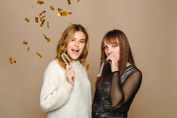 Twee jonge vrouwen op een beige achtergrond in een trui en een zwarte jurk blazen een feestelijk deuntje en houden een suikerriet vast. Kerstmis en Nieuwjaar vieren — Stockfoto