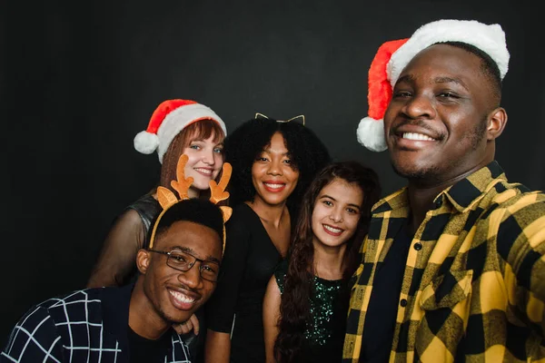 Un grupo de amigos de diferentes nacionalidades se toman un selfie navideño en el estudio. Hombres y mujeres jóvenes en sombreros de santa y cuernos de ciervo se están divirtiendo juntos — Foto de Stock