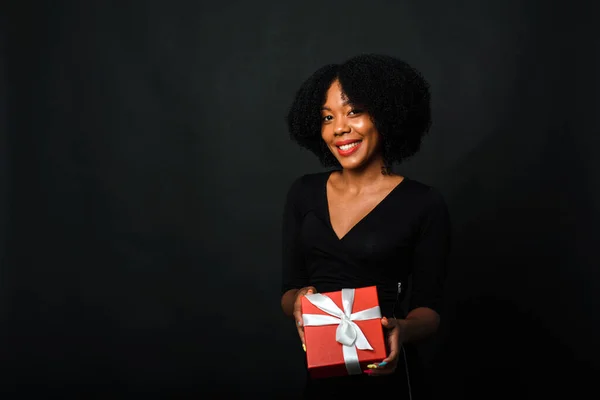 Uma mulher de pele escura com um penteado afro dá um presente para a câmera. O conceito de compras de Ano Novo, presentes. Espaço de cópia — Fotografia de Stock