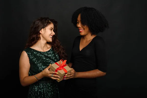 Duas jovens de diferentes nacionalidades estão sorrindo, segurando um presente para o ano novo e o Natal em um fundo preto. Conceito de troca de presentes, dar e receber — Fotografia de Stock