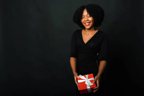 Fekete Péntek. Egy sötét bőrű nő egy újévi ajándékot tart a kezében egy fekete háttérben a stúdióban. — Stock Fotó