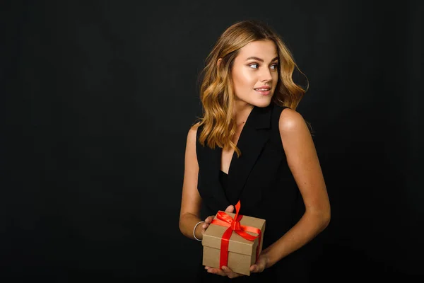 Una hermosa joven sobre un fondo negro sosteniendo un regalo de Navidad con una cinta roja. La felicidad de recibir y dar regalos para el nuevo año — Foto de Stock