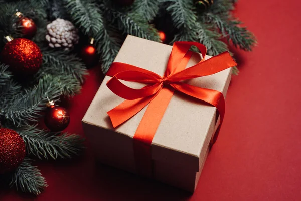 Regalo artesanal con una cinta de satén rojo junto a las ramas y bolas del árbol de Navidad, juguetes sobre un fondo rojo —  Fotos de Stock