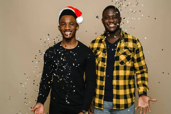 Dois atraentes homens gays negros elegantes estão comemorando a festa de Ano Novo, casal homossexual jogando confete, parabenizando uns aos outros no fundo bege. Celebrando o Natal e Ano Novo — Fotografia de Stock