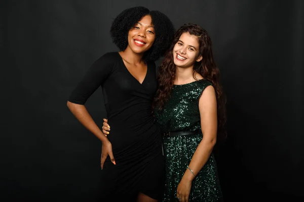 Two lovely young women of different nationalities in beautiful evening dresses stand against a black background, hugging and smiling for the camera — Stock Photo, Image