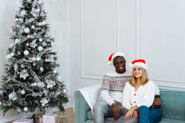 Noel Baba şapkalı çok ırklı genç bir çift bir ağacın yanındaki koltukta oturuyor. Bir adam ve bir kadın kameraya bakıp gülümsüyor.