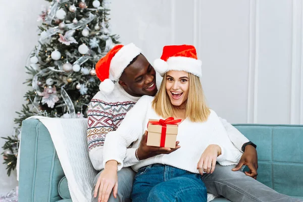 Hombre negro guapo sorprendiendo a una mujer caucásica con un regalo de Navidad. Momentos acogedores para unas vacaciones de invierno. Concepto de Saludos Estacionales. Una mujer se sorprende por el regalo que su marido le dio —  Fotos de Stock
