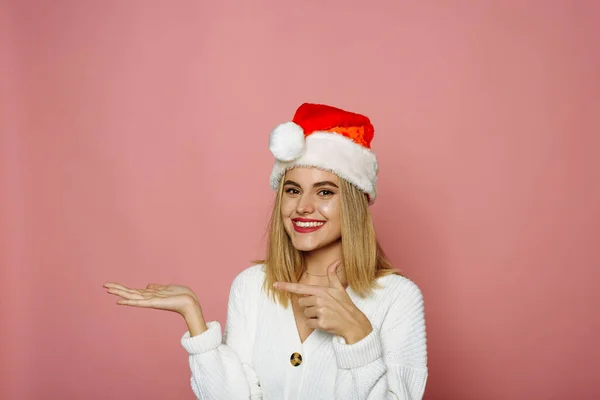 Sonriente joven Santa mujer de 20 años en suéter, en Santa sombrero señalando dedo índice a un lado en el espacio de copia simulada aislado en el fondo rosa, retrato de estudio. Feliz Año Nuevo celebración feliz concepto de vacaciones —  Fotos de Stock