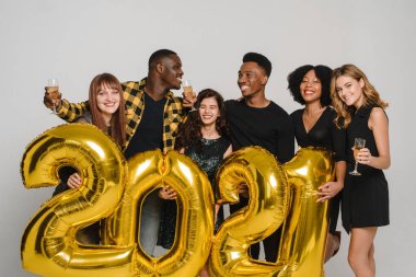 Neşeli arkadaşlar 2021 numarayı tutuyor ve yeni yılı kutluyorlar. - Şerefe. Bir grup çok ırklı arkadaş şampanya içiyor ve beyaz bir arka planda eğleniyorlar.