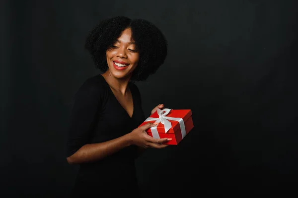 Egy fekete nő ünnepi fekete ruhában egy karácsonyi piros ajándékot tart fehér szalaggal a kezében. Az ajándékozás vagy fogadás koncepciója az új évre — Stock Fotó