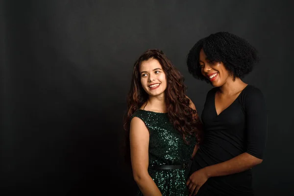 Twee mooie jonge vrouwen in mooie avondjurken staan op een zwarte achtergrond, knuffelen en lachen — Stockfoto