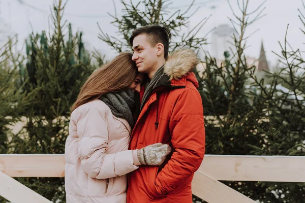 Beautiful young couple stand face to face among christmas tree. Young newlyweds in love, gently kissing, hugging, dressed in warm clothes, snow-covered city background, love story