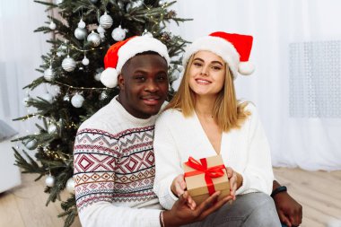 Birbirine aşık bir çift Noel için dekore edilmiş oturma odasında oturuyor ve hediyelerini tutuyor. İkisi de kameraya mutlu bir şekilde bakıyor. Mutlu Noeller ve mutlu yıllar.