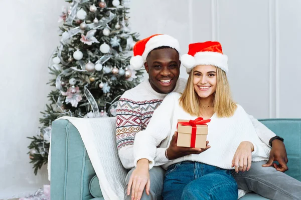 Joven feliz pareja multiétnica abrazándose y relajándose en el cómodo sofá. Joven hombre y mujer están mirando a la cámara y sonríen. Retrato de pareja encantadora en época de Navidad —  Fotos de Stock