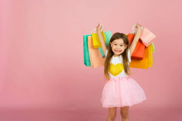 Kleine blonde Mädchen genießt ihren Einkauf auf pastellrosa Hintergrund mit Copyspace. Verkauf. Nettes kleines Mädchen mit vielen bunten Einkaufstaschen im Atelier — Stockfoto