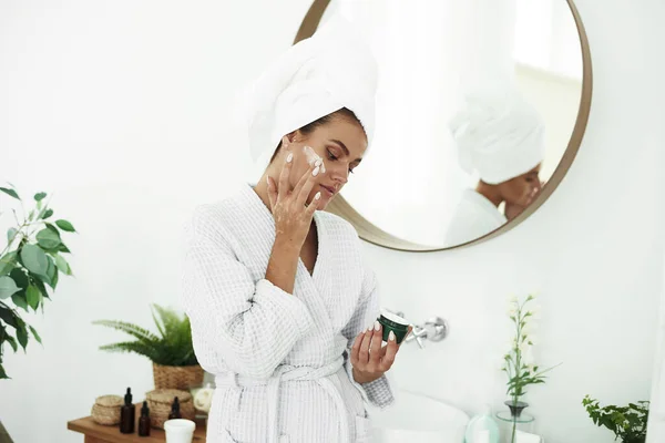Retrato de una joven sonriente aplicándose crema hidratante en la cara en el baño. Cosmetología. Belleza y Spa — Foto de Stock