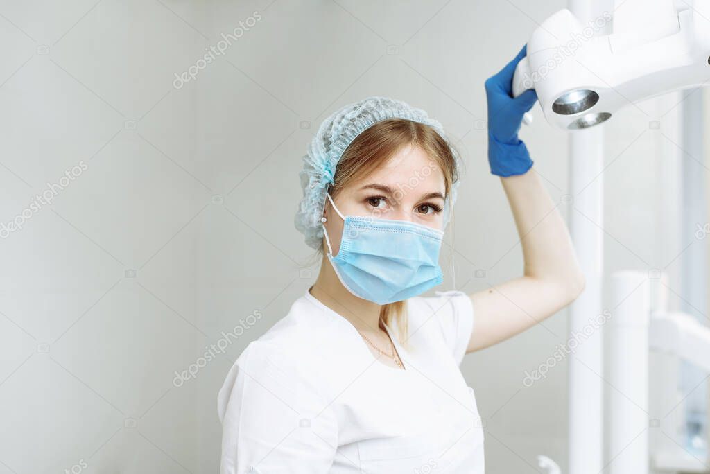 Portrait of young female dentist wearing surgical mask examines a patient while holding dental lamp