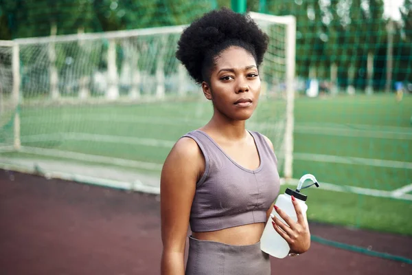 Jovem mulher negra em um traje de treino fica no chão de esportes e mantém uma garrafa de água. Conceito de estilo de vida saudável. Ao ar livre — Fotografia de Stock
