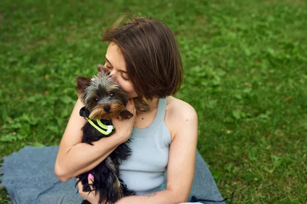 Милая девушка с квадратными объятиями обнимает йоркширского терьера на улице. Обнимается с маленькой собачкой в парке — стоковое фото