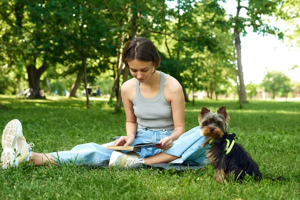 Молодая женщина читает книгу на природе рядом со своим маленьким псом Йорширом Терьером — стоковое фото