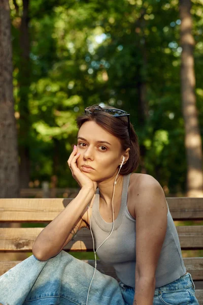 Mujer joven con estilo de moda escuchando música en auriculares con cable mientras está sentado en un banco del parque — Foto de Stock