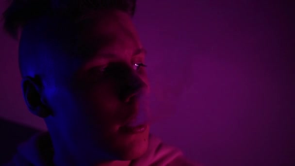 En ung kille i luvtröja andas ut rök i neonljus. Tonåringen röker på klubben. Närbild — Stockvideo