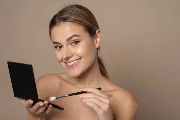 A lovely young woman applies brown eyeshadow to her eyes from an eyeshadow palette. Studio portrait of a woman who does everyday makeup