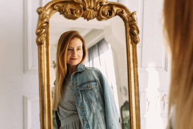 Sıcak ev atmosferi. Gri elbiseli ve kot pantolonlu şık kızıl kadın evde bir sandalyeye oturup aynaya bakıyor. Portre güzel genç kadın mutlu gülümseme oturma odasında rahatla.