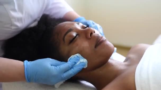 Очистка кожи хлопчатобумажными прокладками в офисе косметолога. Темнокожая молодая женщина лежит на процедурах у косметолога. Уход за кожей, очищение и увлажнение, спа-концепция — стоковое видео