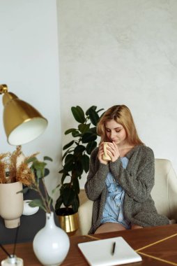 A cute young woman makes notes in a notebook while sitting at her desk at home and drinking coffee