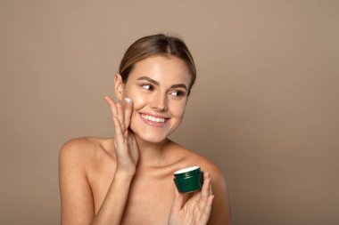 Young woman applies moisturizer to her face and smiles. Skin care concept. clipart