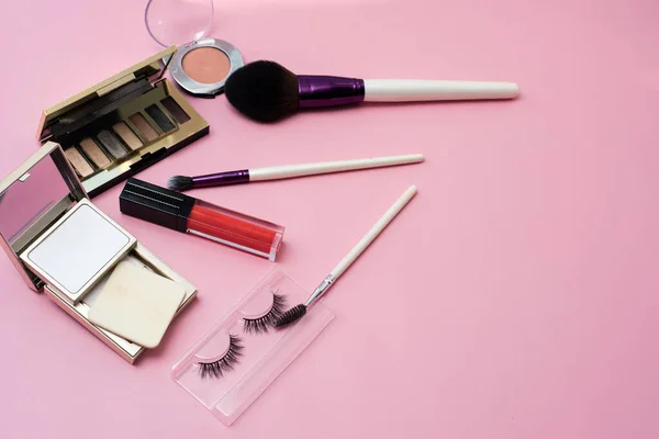 Dekorativ kosmetika på en rosa bakgrund. Läppglans, sminkborstar, puder, ögonskugga och lösögonfransar för ljus kvinnlig makeup. Kopiera utrymme — Stockfoto