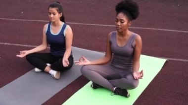 Dışarıda yoga yapan iki güzel kadın. Beyaz ve siyah kadın stadyumda lotus pozisyonunda oturuyorlar.