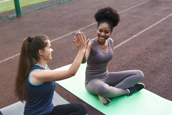 Två kvinnliga idrottare high-five sittande på en yogamatta utomhus. Lagarbete i idrottsträning — Stockfoto