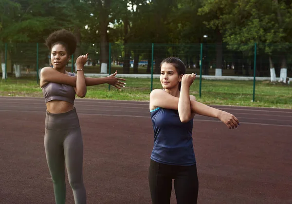 Två kvinnor av olika nationaliteter värms upp innan de tränar utomhus. Uppvärmning av muskler före styrketräning eller jogging — Stockfoto