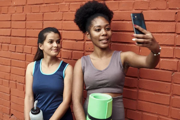 Elképesztő fiatal atlétikai nők képe, amint szelfit készítenek egy téglafalnak. Mosolygó fekete nők időt töltenek együtt edzés után — Stock Fotó