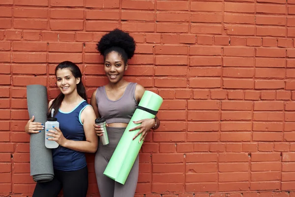 Två vackra unga kvinnor i träningsoveraller står mot en tegelvägg på gatan. Flickvännerna ler medan de håller yogamattor och vattenflaskor — Stockfoto