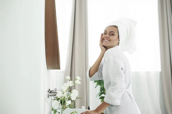 Eine hübsche junge Frau im Bademantel und mit einem Handtuch umwickeltem Haar blickt nach der Dusche in den Spiegel. Pflege-, Reinigungs- und Feuchtigkeitskonzept der Haut — Stockfoto