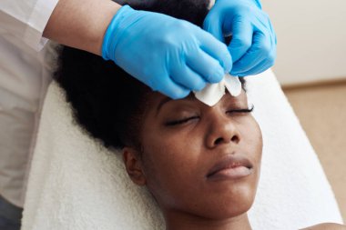 Kozmetik uzmanı, güzellik salonundaki genç ve güzel bir kadının derisini tonikle temizler. Koyu tenli model güzellik uzmanının kanepesinde yatıyor.