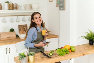 Portakal suyu içen mutlu genç bir kadın yemek pişirirken sosyal ağlara göz atıyor. Ev hanımı kameraya bakıp gülümsüyor.